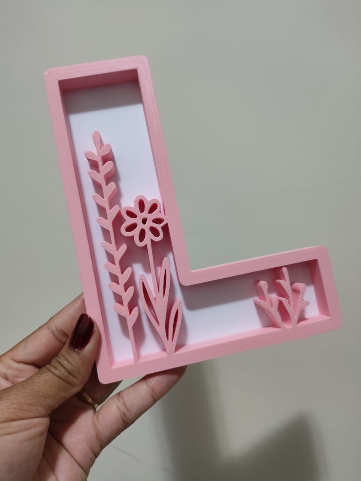 Letra L decorativa em rosa blush com motivos florais no interior sobre fundo branco, segurada por uma mão.