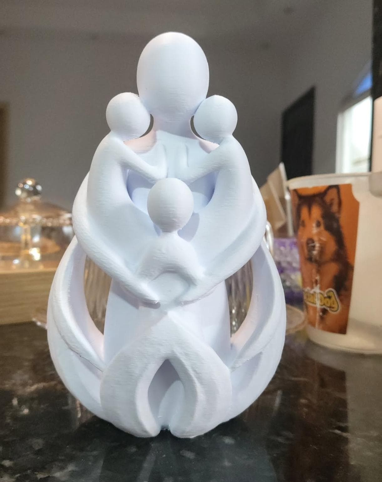 Escultura branca minimalista com figuras abraçadas sobre tampo escuro, fundo doméstico desfocado.