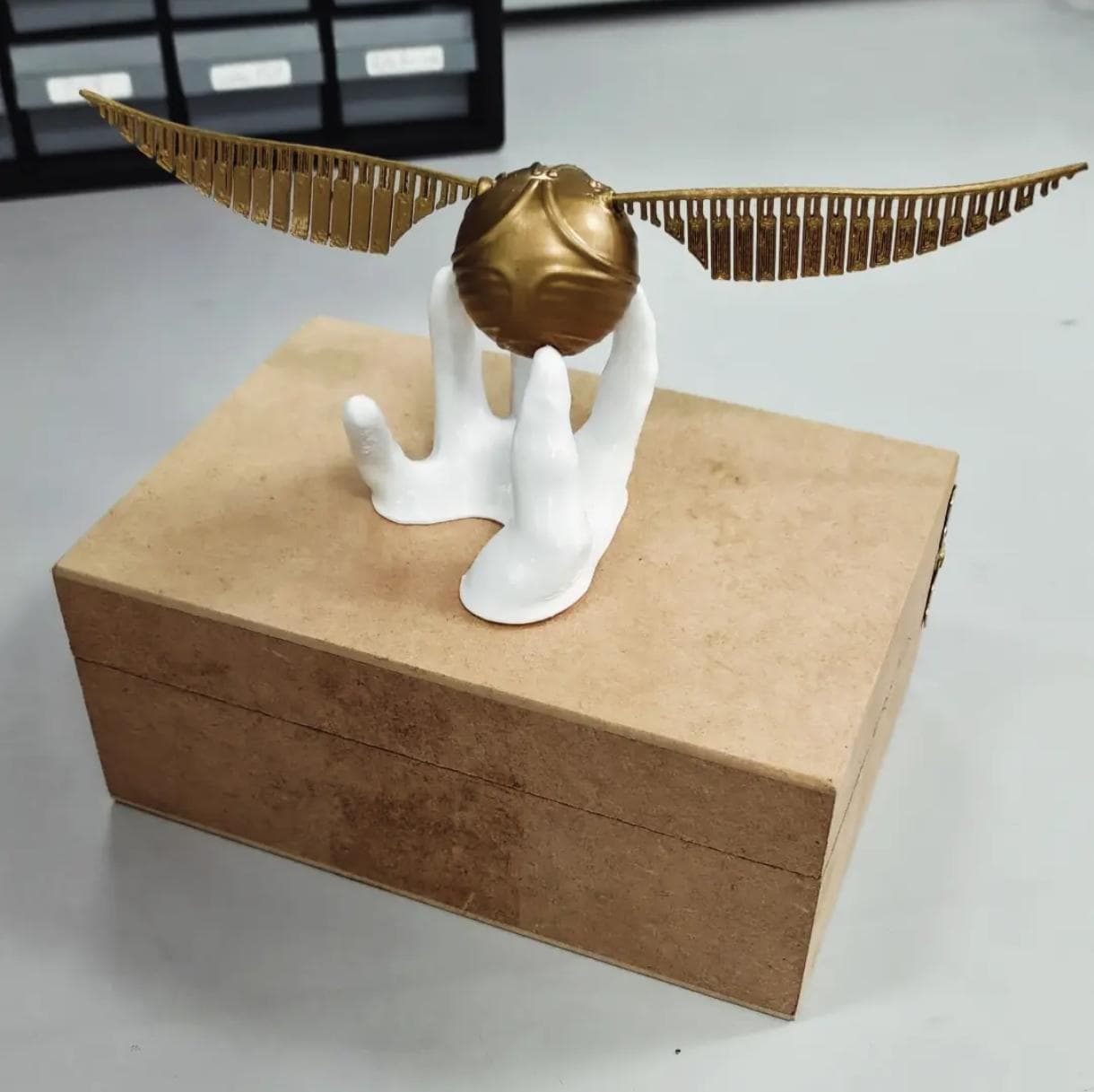 Pomo dourado com asas sobre suporte branco em forma de mão, sobre caixa de madeira MDF.